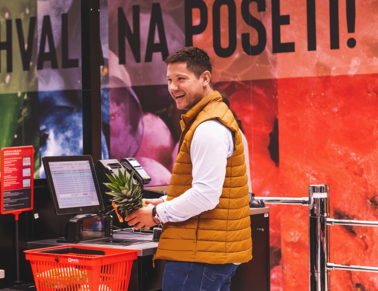 A customer from Maxi is standing in front of the self scan with a pineapple while he is smiling.